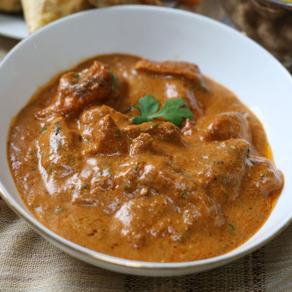 A white bowl filled with rich, orange-brown Indian curry, garnished with a green herb.