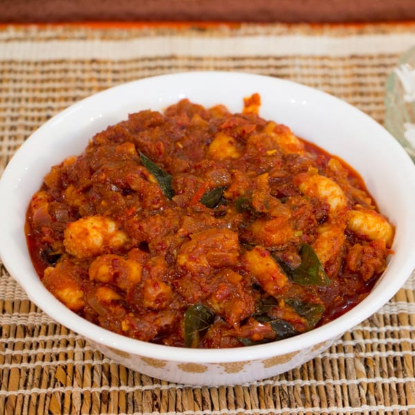 A white bowl filled with spicy red Indian shrimp curry, garnished with curry leaves, on a woven placemat.